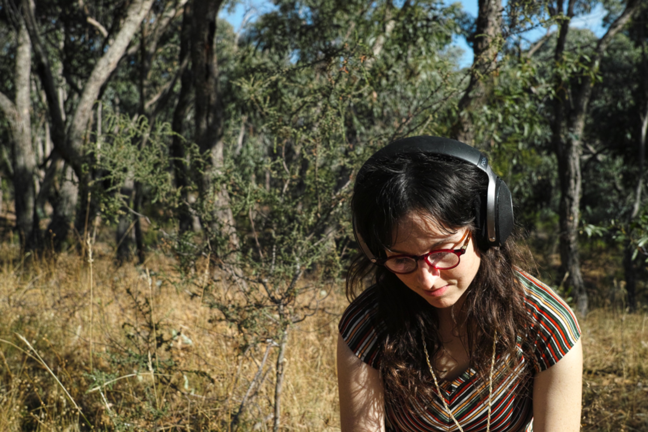 Emily Kostos, a woman with long dark hair and red-framed glasses, wears large over-ear headphones while standing in a dry, wooded landscape. She is dressed in a striped, short-sleeved top and appears focused, looking downward. Sunlight filters through the sparse eucalyptus trees, casting dappled shadows on the ground. Photo credit: Rowan Chase.
