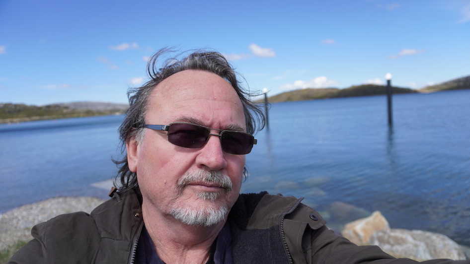 Photo of Julian Knowles, head shot with Rocky Valley dam in background with distant mountains. Blue skies and white clouds.