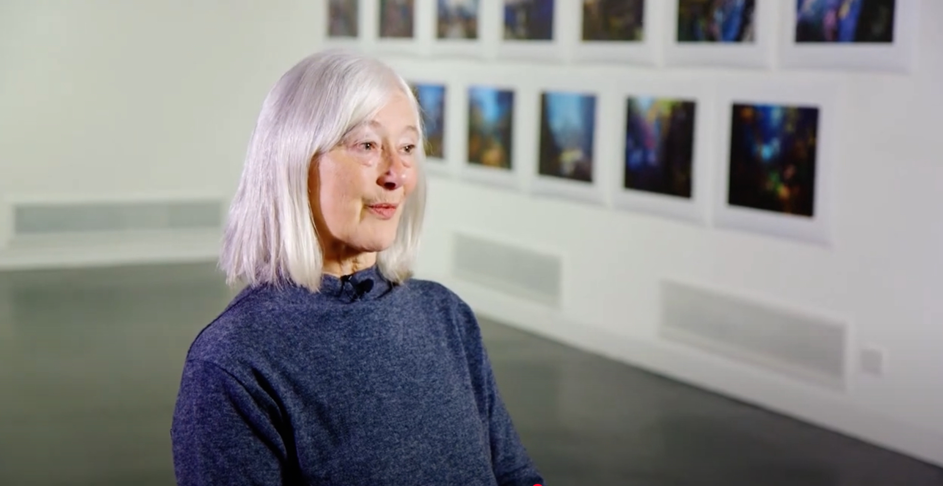 Photo of Lesley Duxbury sitting in a gallery, speaking about her artwork. Multiple blurred images are visible behind her.